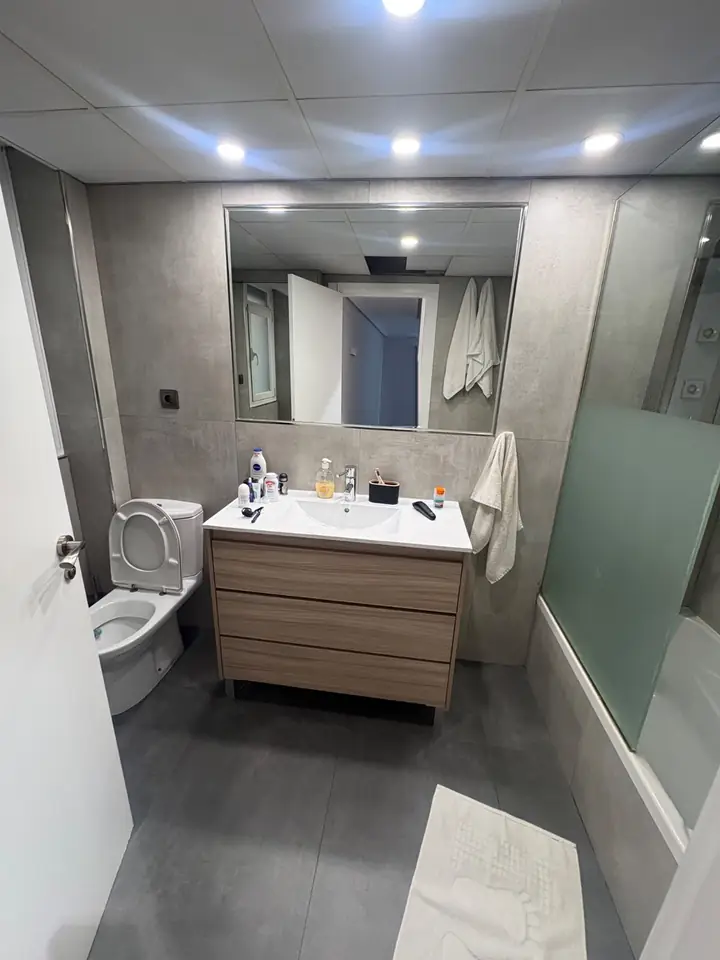 Foto real de un baño antes de la remodelación: lavamanos con mueble de madera, sanitario, tina con división de baño y cerámica gris